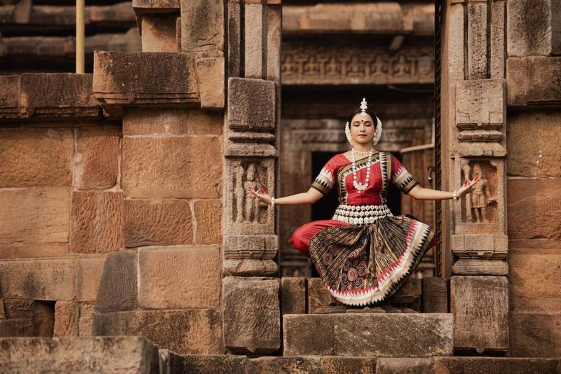 Seated in chauka within a carved temple doorway, arms extended sideways in mudra, eyes lowered.
