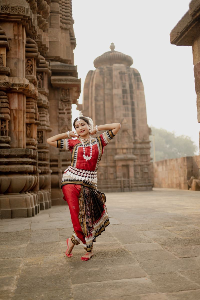 Yaman Kalyan Pattanaik in red Odissi costume, both arms lifted to the head, mid-phrase on the stone courtyard of a temple in Odisha.