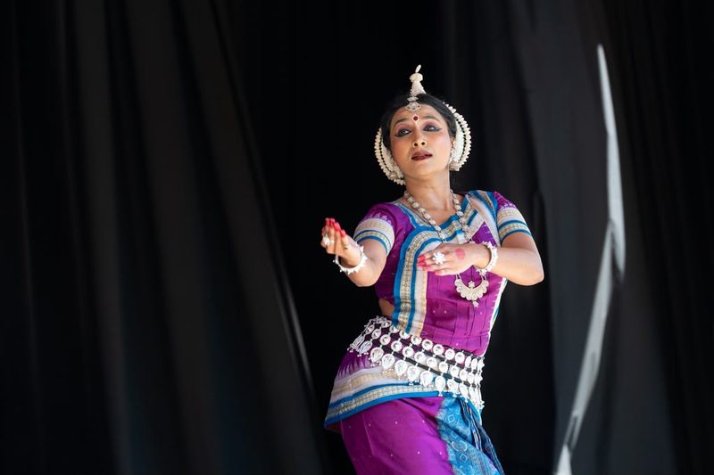 Yaman in purple and gold Odissi costume, both hands held in mudra mid-phrase, against a black stage backdrop.