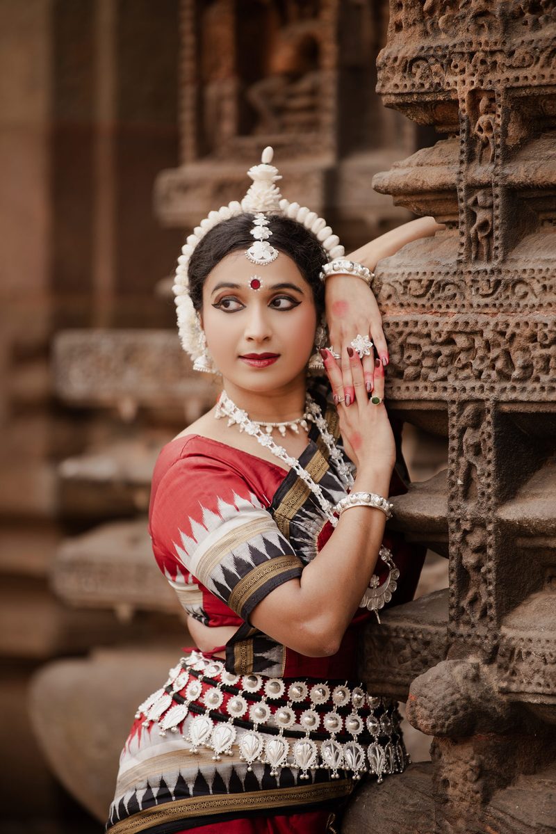 Yaman in red and black Odissi costume resting one arm against a carved temple pillar, looking over her shoulder.