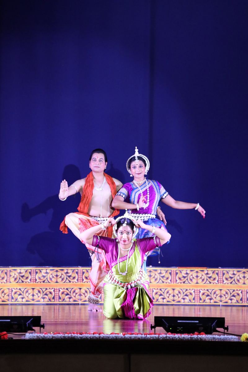 Three Odissi dancers holding a layered group composition on a blue-lit stage.