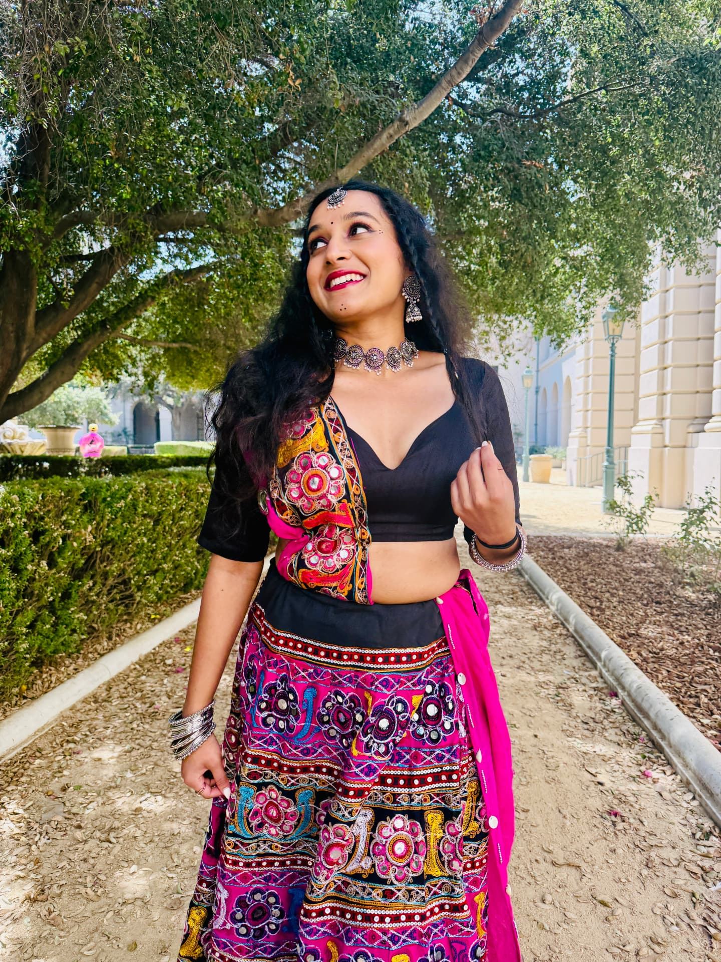 Yaman smiling in a colourful mirrorwork costume and silver jewellery, standing under a tree.