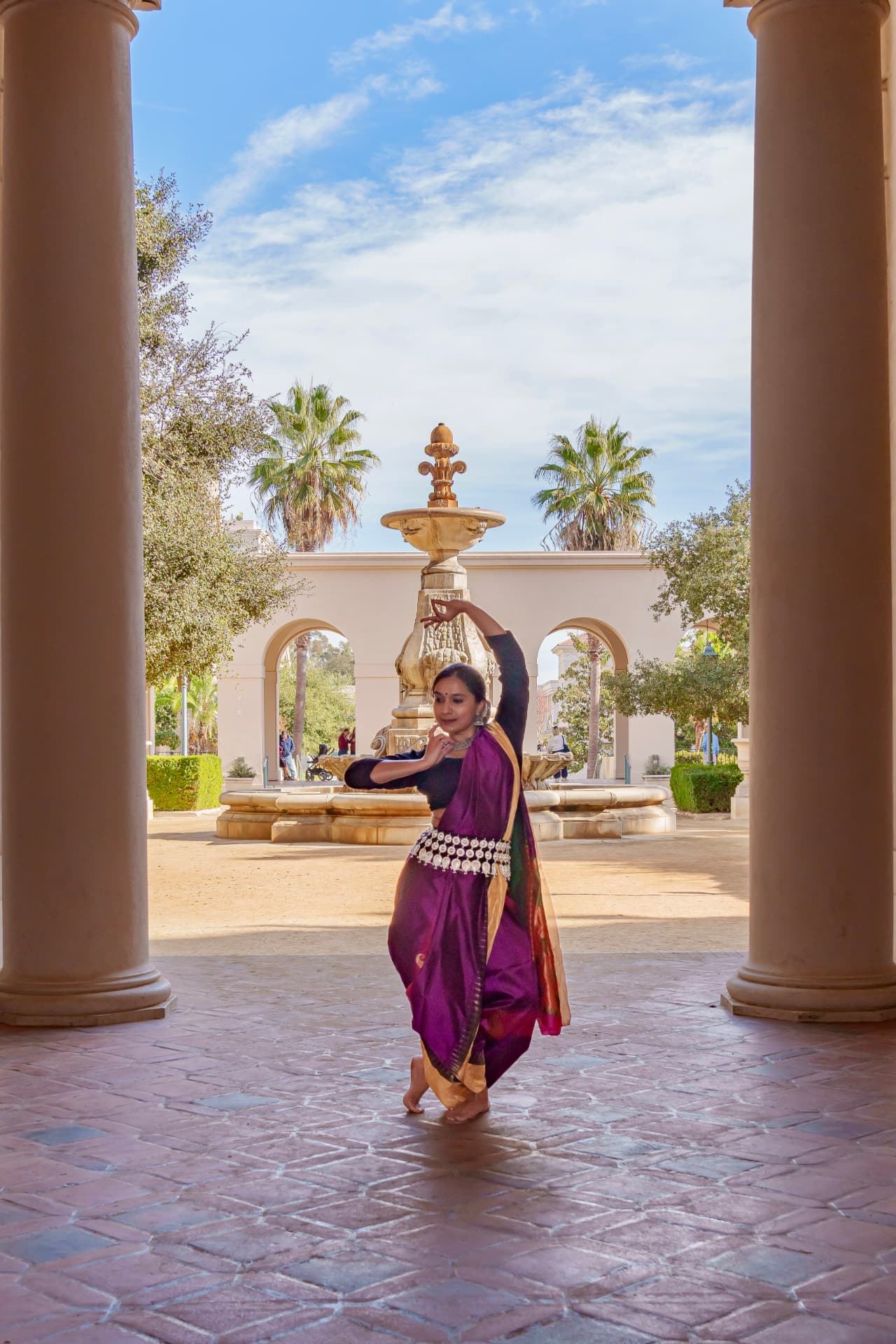 Dancing in a purple sari between columns with a fountain in the background, one arm raised high.