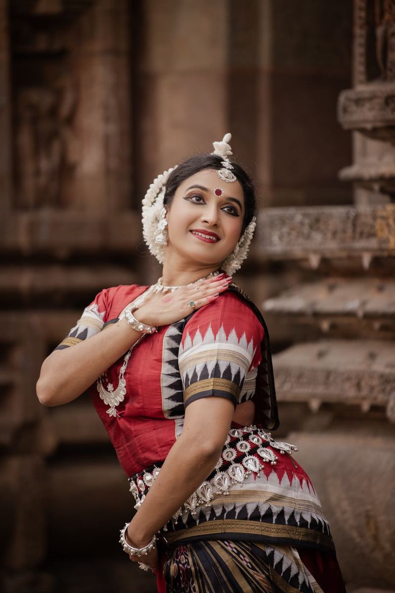 Turning over one shoulder in red Odissi costume with silver jewellery, one hand resting at the collarbone.