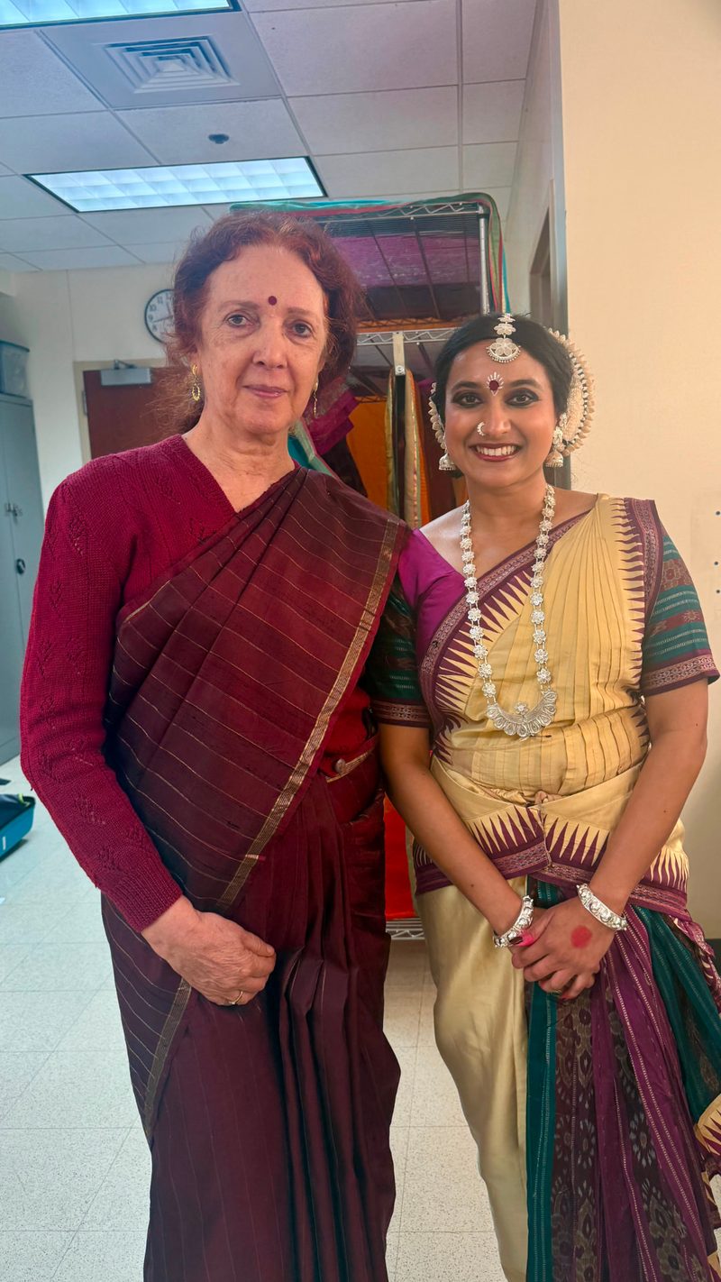 Yaman in cream and maroon Odissi costume with full silver jewellery, standing backstage beside her guru after a performance.