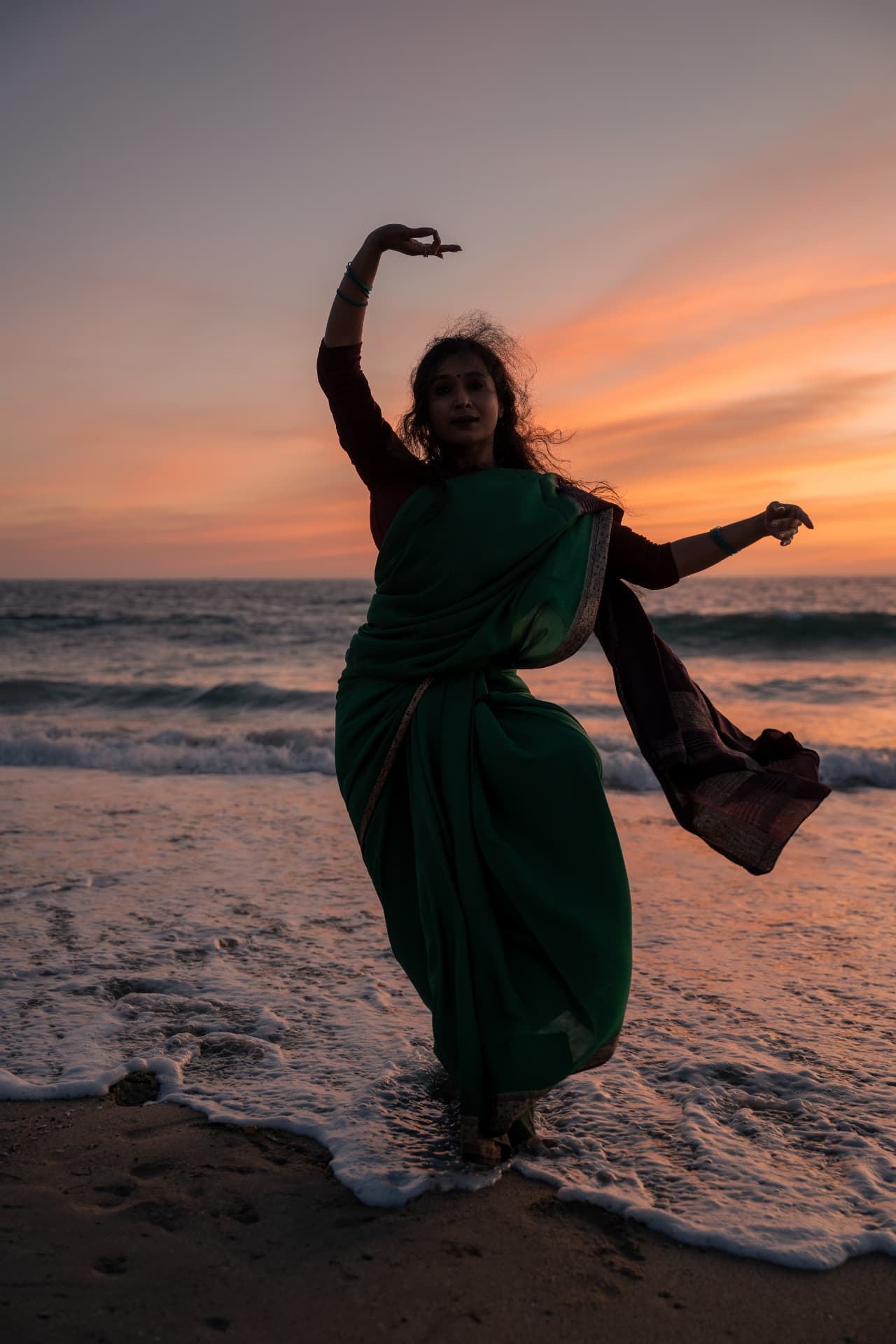 Dancing in a green sari at the edge of the surf, sunset colours behind.