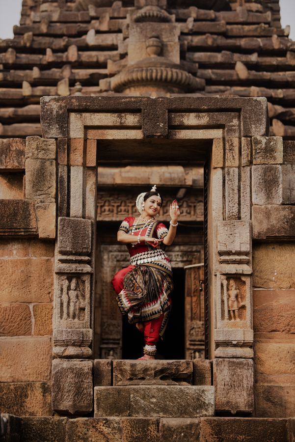 Standing on one leg inside a temple doorway framed by carved sandstone, one hand raised in abhaya.