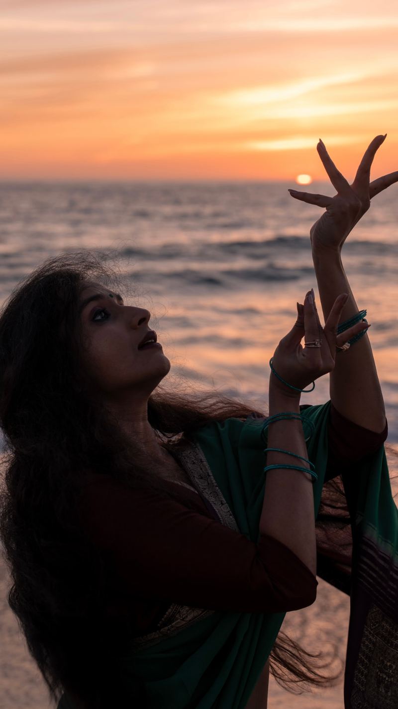 Detail of hands held in mudra against a sunset sky over the ocean.