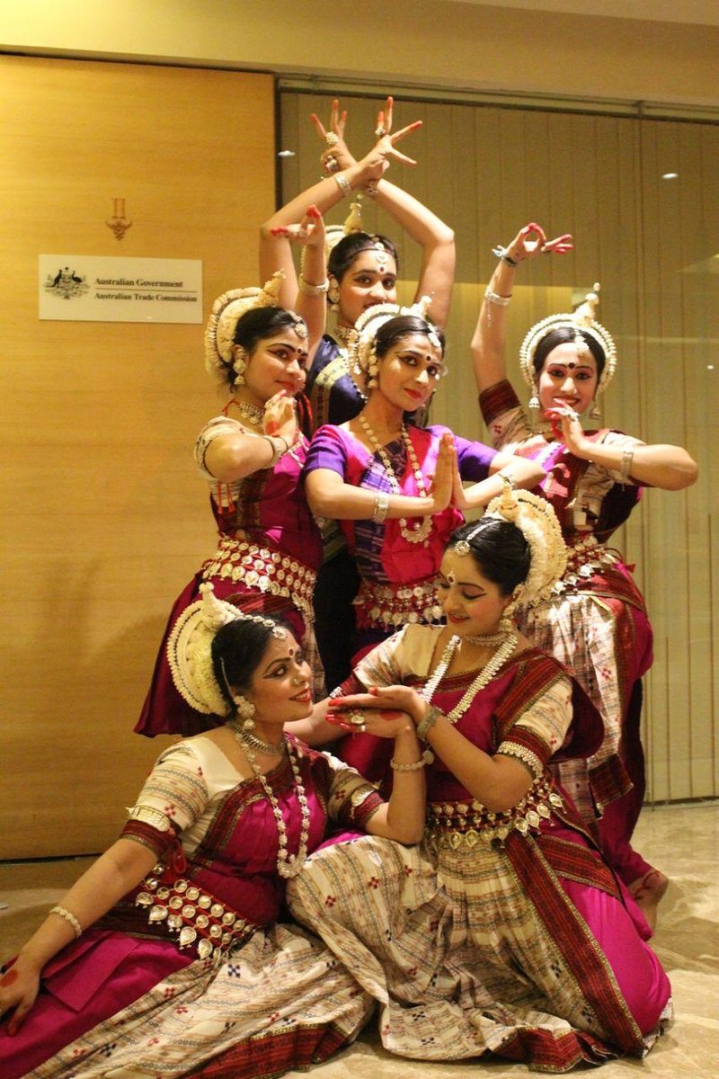 Six Odissi dancers in magenta and cream costumes holding a layered group tableau, hands in mudras, at an indoor venue.