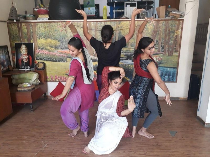 Four dancers rehearsing barefoot in a home studio, arms raised mid-phrase in front of a painted backdrop.