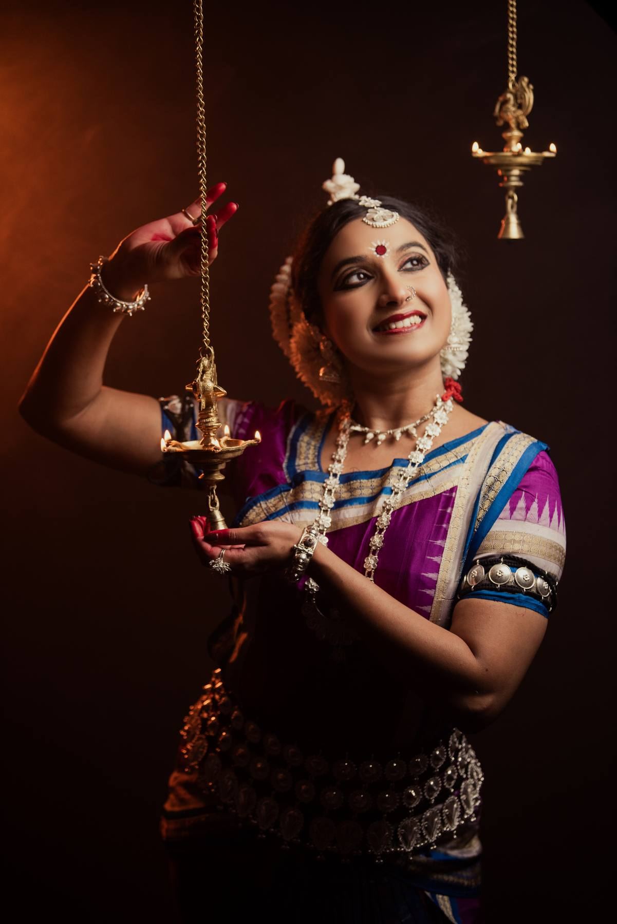 Holding a lit brass oil lamp on its chain, smiling upward, in purple and blue silk against a dark studio ground.