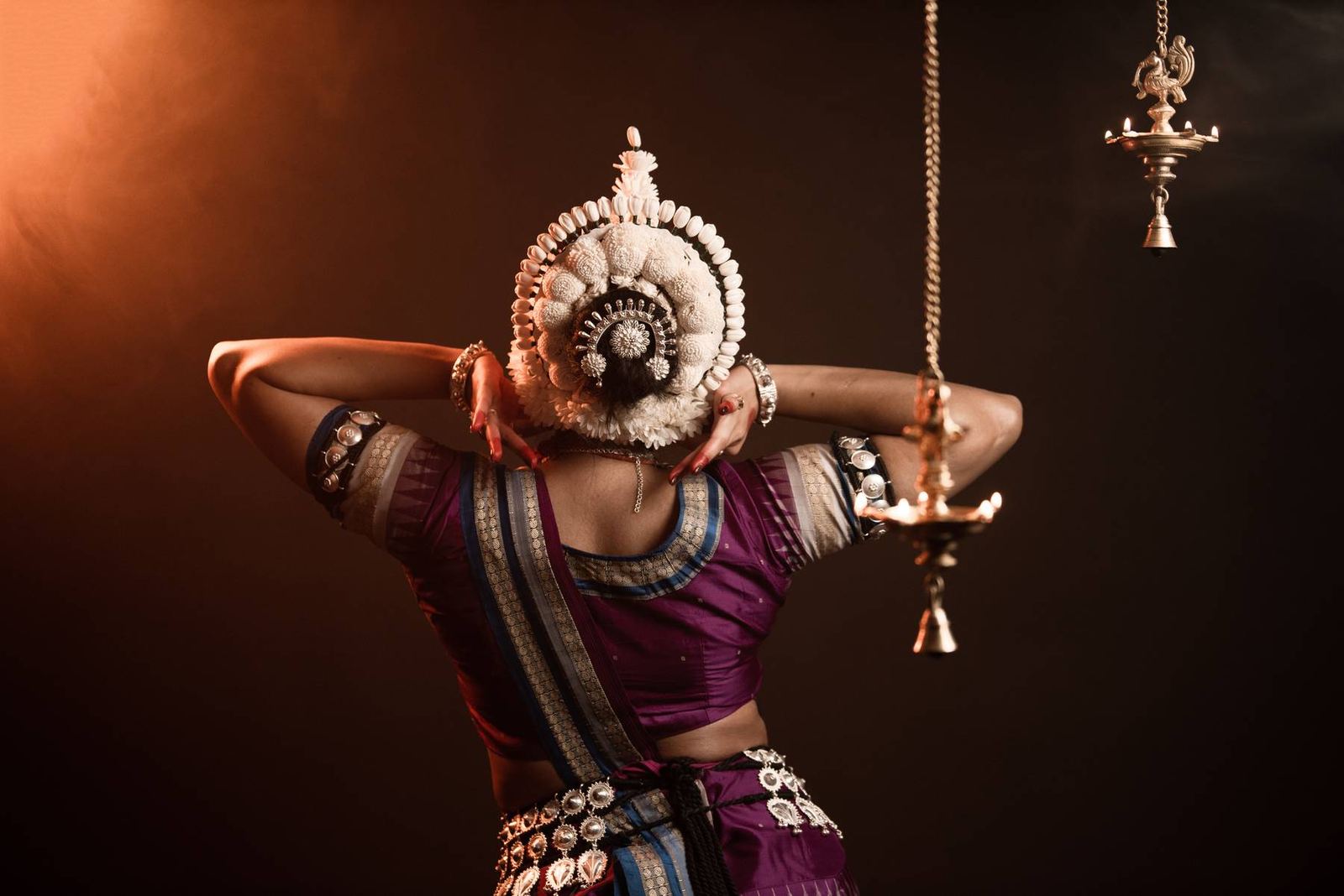 Seen from behind in purple silk with a shell headdress, arms raised behind the head, brass oil lamps burning to one side.