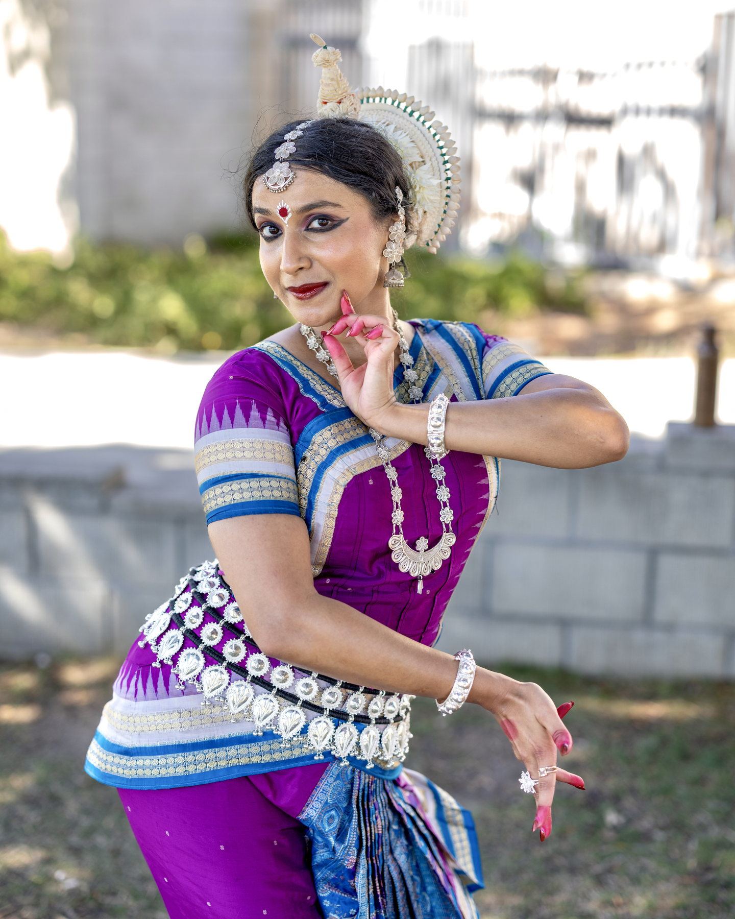 Outdoor portrait in purple and blue Odissi silk with shell headdress, one hand raised in mudra near the chin.
