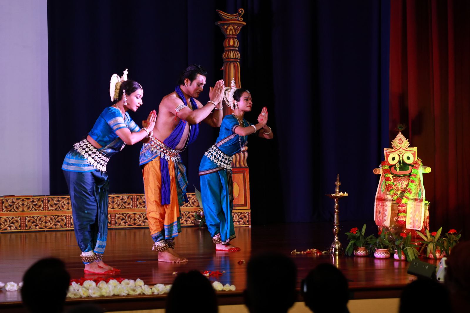 Three Odissi dancers bowing in namaskar on a lamp-lit stage beside a Jagannath icon.