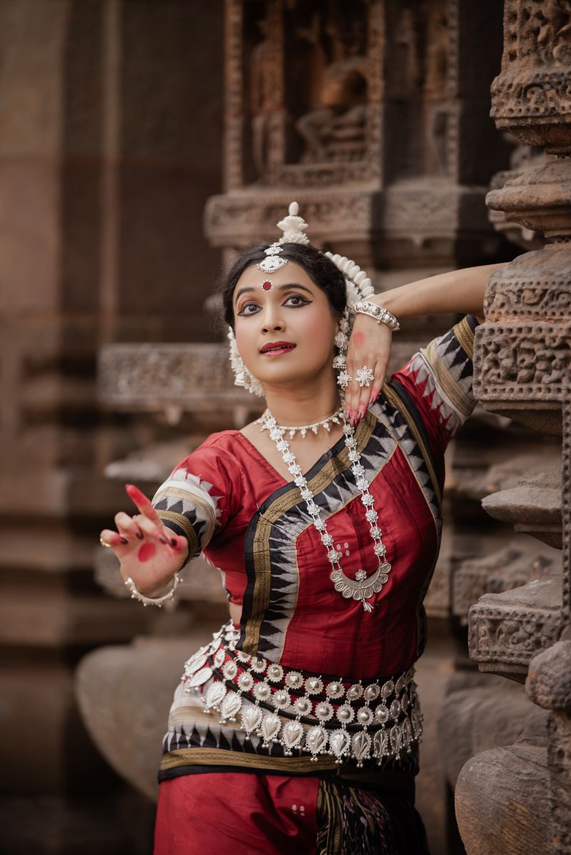 Leaning against a carved temple pillar in red silk, one hand extended forward in mudra, looking past the camera.