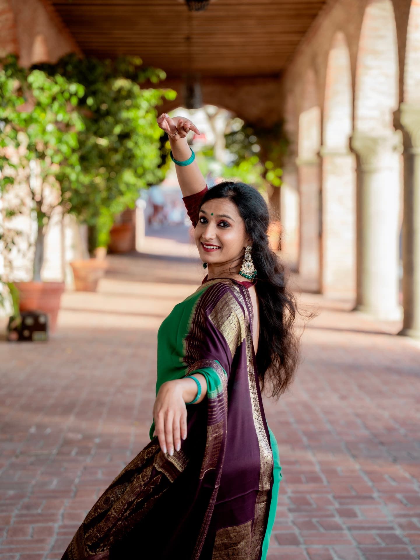 Yaman Kalyan Pattanaik in a green and plum silk sari, posing in a brick corridor with one arm raised in mudra.