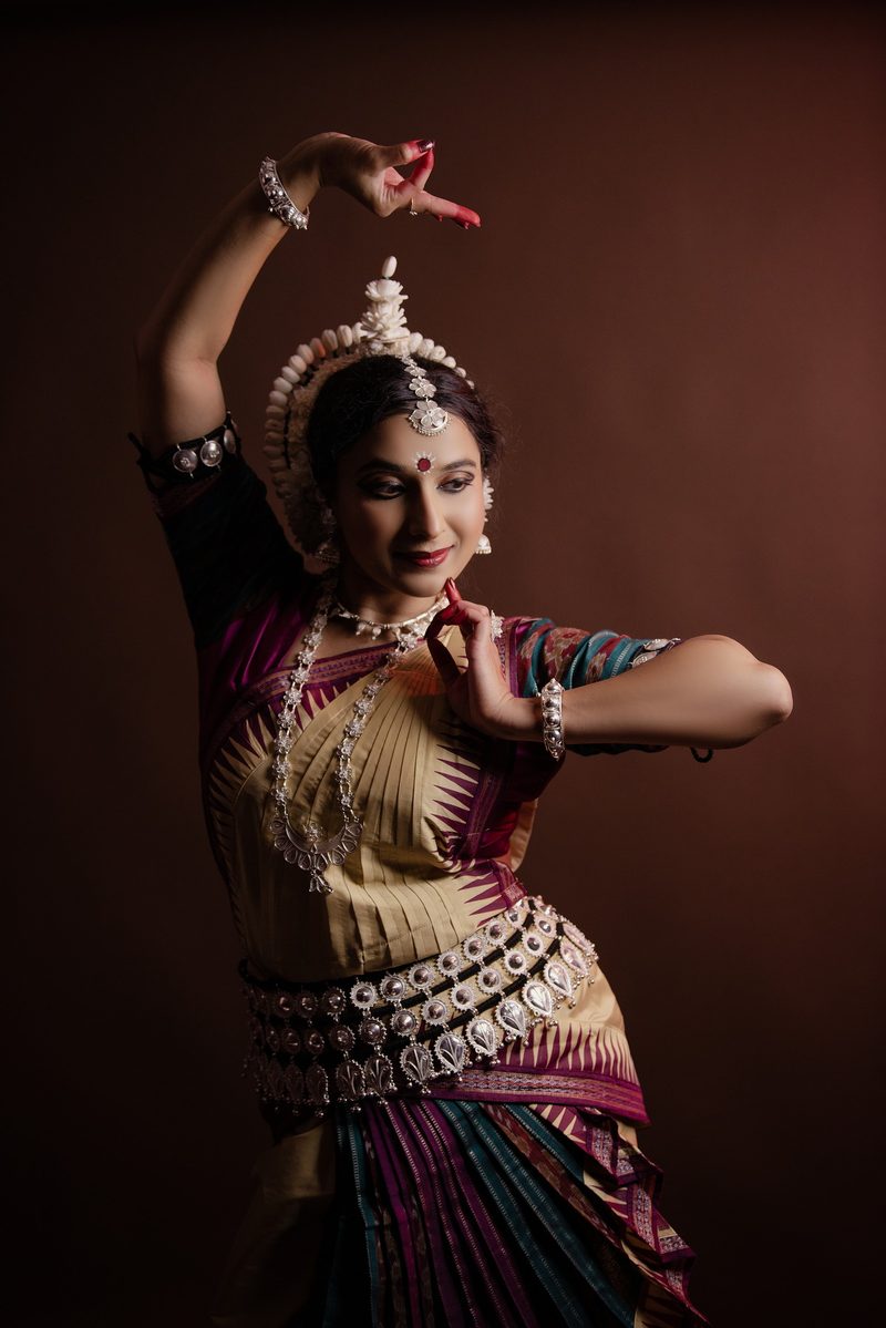 Studio portrait in cream and plum silk, one arm arched overhead and the other hand in mudra at the chin.