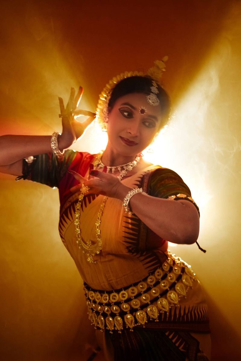 Studio portrait backlit in gold, both hands held in mudra close to the face, gaze turned down and to the side.