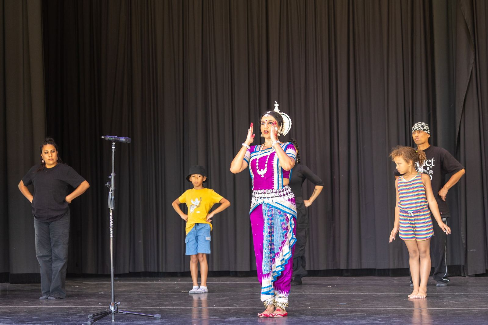 Yaman in magenta Odissi costume calling out mid-performance, flanked by community dancers on a festival stage.