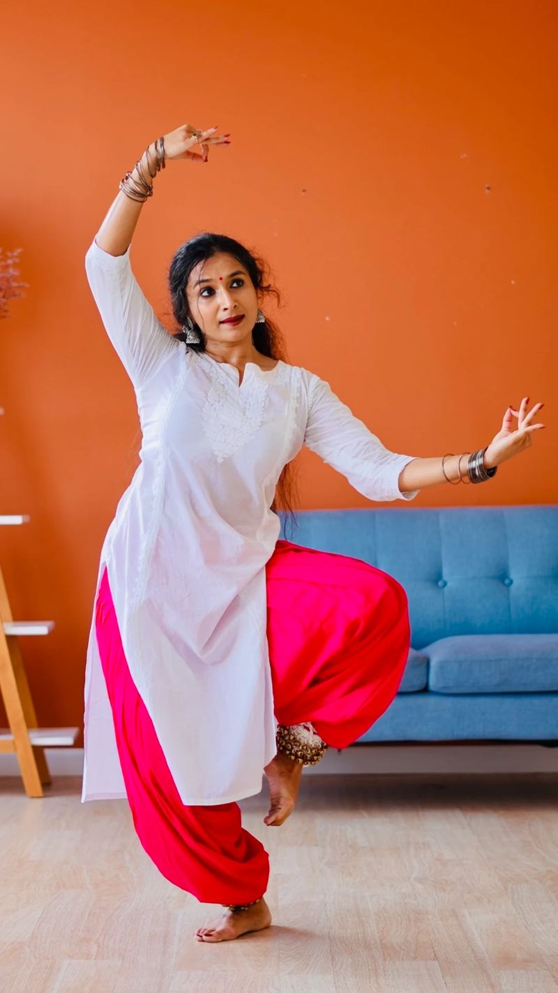 Yaman in a white and pink outfit balanced on one leg indoors against a warm orange wall.