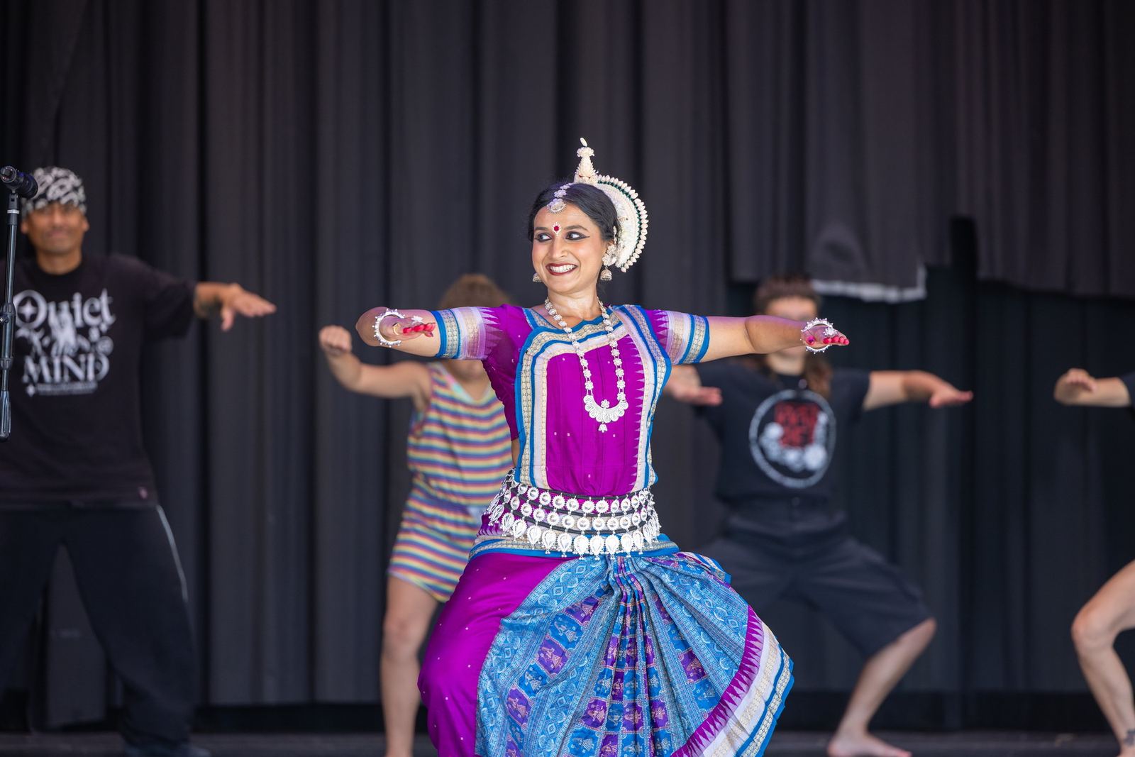 Yaman smiling mid-phrase in magenta Odissi costume, arms extended wide, with other dancers behind her on an outdoor stage.